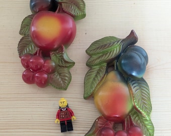 May include: Two decorative wall plaques shaped like fruit clusters. One features a large red apple, a dark plum, and red berries with green leaves. The other shows a pear, a dark plum, and red berries with green leaves. A small yellow Lego figure.