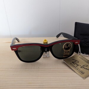 May include: Black Ray-Ban sunglasses with red accents on the top frame and green lenses. The sunglasses are displayed with a black case and a tag that reads "Ray-Ban Bausch & Lomb".