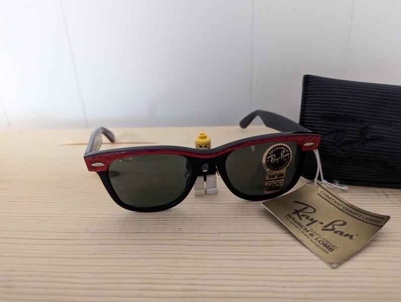 May include: Black Ray-Ban sunglasses with red accents on the top frame and green lenses. The sunglasses are displayed with a black case and a tag that reads "Ray-Ban Bausch & Lomb".