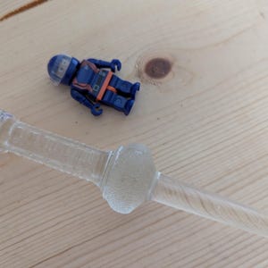 May include: A clear glass bottle stopper with a textured, bulbous center and a long, slender stem. A small blue and orange action figure is next to the stopper on a light wood surface. The stopper is likely for a wine bottle.