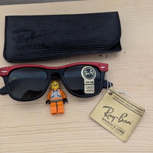 May include: Black Ray-Ban Wayfarer sunglasses with red accents on the top frame, a black case, and a tag. A small Lego figure is placed near the sunglasses. The tag reads "Ray-Ban Bausch & Lomb".