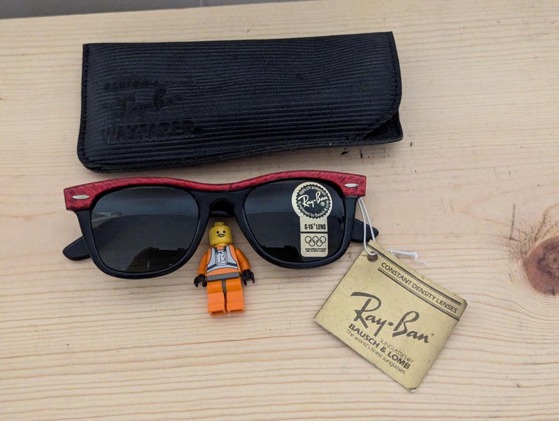 May include: Black Ray-Ban Wayfarer sunglasses with red accents on the top frame, a black case, and a tag. A small Lego figure is placed near the sunglasses. The tag reads "Ray-Ban Bausch & Lomb".
