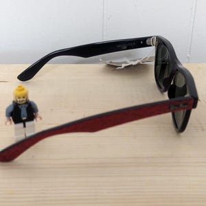 May include: Black Wayfarer sunglasses with red patterned arms. The sunglasses are on a light wood surface. A small Lego figure is in the foreground.