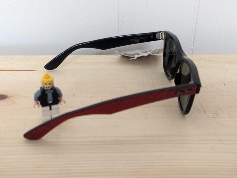 May include: Black Wayfarer sunglasses with red patterned arms. The sunglasses are on a light wood surface. A small Lego figure is in the foreground.