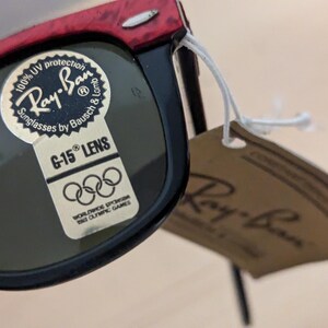 May include: Black and red Ray-Ban sunglasses with a G-15 lens. The sunglasses have a tag attached with the Ray-Ban logo and text that reads "100% UV protection Sunglasses by Bausch & Lomb". The tag also includes the text "Worldwide Sponsor 1962 Olympic Games".