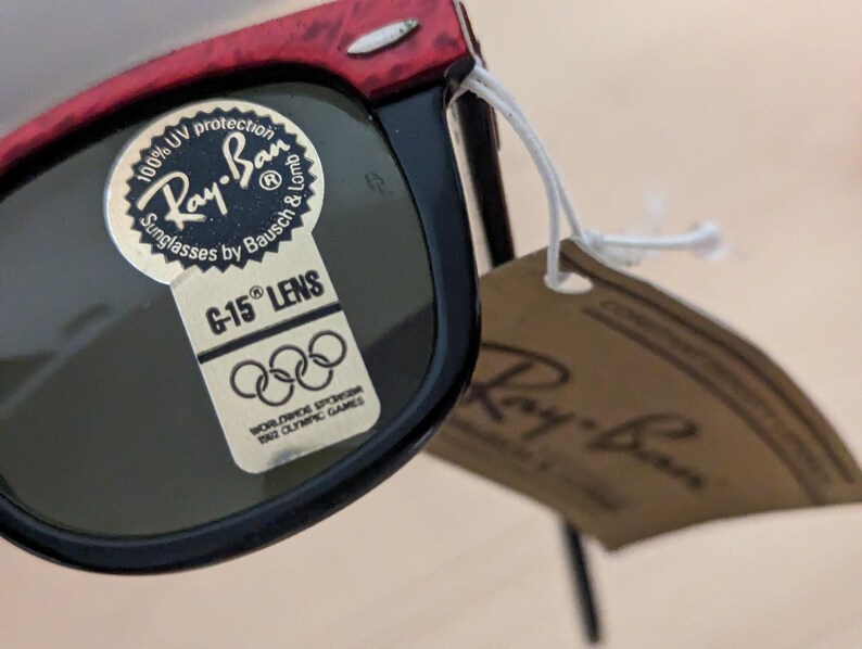 May include: Black and red Ray-Ban sunglasses with a G-15 lens. The sunglasses have a tag attached with the Ray-Ban logo and text that reads "100% UV protection Sunglasses by Bausch & Lomb". The tag also includes the text "Worldwide Sponsor 1962 Olympic Games".