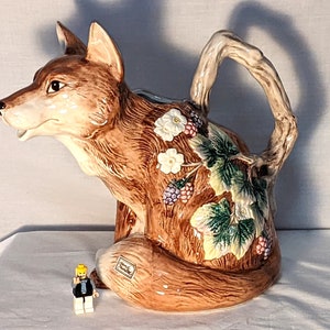 May include: A ceramic fox-shaped teapot with a branch-like handle. The teapot is brown with white flowers, green leaves, and purple berries. A small Lego figure is in front of the teapot.