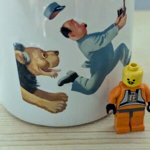 May include: White ceramic mug with a whimsical illustration of a man, a dog, and a Lego figure. The man is depicted in mid-air, and the dog is emerging from the side of the mug. A small orange Lego figure stands in front of the mug.