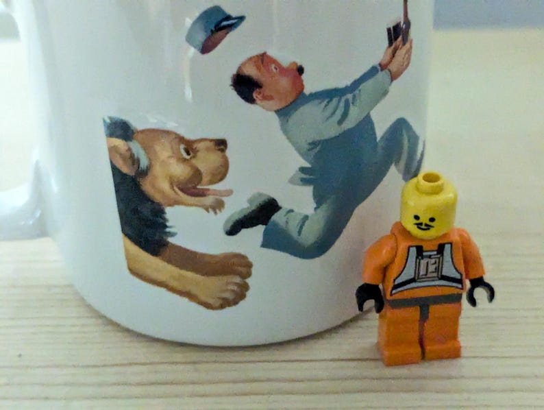 May include: White ceramic mug with a whimsical illustration of a man, a dog, and a Lego figure. The man is depicted in mid-air, and the dog is emerging from the side of the mug. A small orange Lego figure stands in front of the mug.