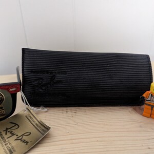 May include: Black Ray-Ban sunglasses with a red accent on the frame, a black case, and a tag. The tag reads "Ray-Ban" and "BAUSCH & LOMB". A small Lego figure in an orange uniform stands next to the case.
