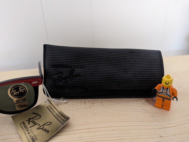 May include: Black Ray-Ban sunglasses with a red accent on the frame, a black case, and a tag. The tag reads "Ray-Ban" and "BAUSCH & LOMB". A small Lego figure in an orange uniform stands next to the case.