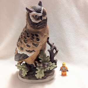 May include: A ceramic owl figurine featuring brown, tan, and white feathers, perched on a dark branch with green oak leaves and acorns. A small orange and gray plastic toy figure stands next to the base to provide a sense of scale for the owl.