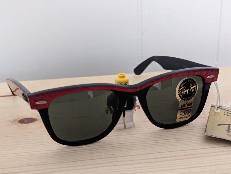 May include: Black sunglasses with a red patterned top frame and dark lenses. The sunglasses have a Ray-Ban logo sticker and a tag attached. The sunglasses are on a wooden surface.
