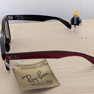 May include: Black Ray-Ban sunglasses with a red patterned arm and a tag. The tag reads "CONSTANT DENSITY LENSES" and "Ray-Ban". A small Lego figure stands near the glasses.