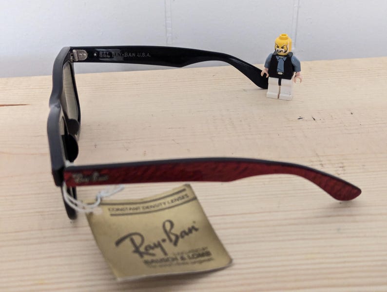 May include: Black Ray-Ban sunglasses with a red patterned arm and a tag. The tag reads "CONSTANT DENSITY LENSES" and "Ray-Ban". A small Lego figure stands near the glasses.