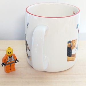 May include: White ceramic mug with a red rim, featuring colorful illustrations of various characters. A small orange and black Lego figure stands beside the mug on a light wood surface. The mug has a large white handle.