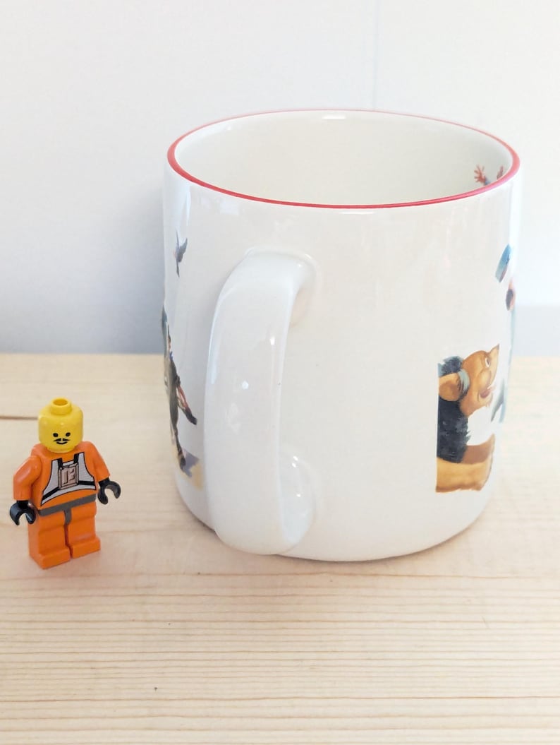May include: White ceramic mug with a red rim, featuring colorful illustrations of various characters. A small orange and black Lego figure stands beside the mug on a light wood surface. The mug has a large white handle.