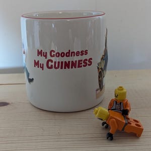 May include: White ceramic mug with red trim and the text "My Goodness My Guinness" in red. A small Lego figure in an orange jumpsuit is riding a Lego brick. The mug has illustrations of people and vehicles.