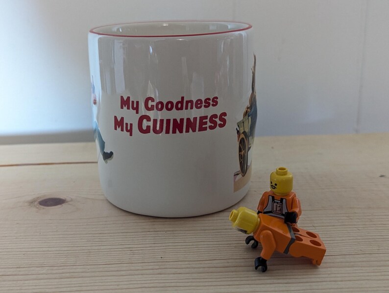 May include: White ceramic mug with red trim and the text "My Goodness My Guinness" in red. A small Lego figure in an orange jumpsuit is riding a Lego brick. The mug has illustrations of people and vehicles.