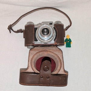 May include: A vintage Edinex camera with a silver body and black accents, housed in a brown leather case with a matching shoulder strap. A small yellow and green plastic toy figure stands next to the camera for scale, providing a size reference.