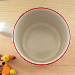 May include: A white ceramic mug with a red rim and handle. The mug is empty and sits on a light brown wooden surface. Two small toy figures are next to the mug.