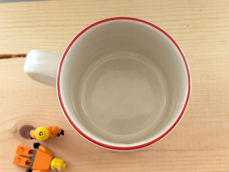 May include: A white ceramic mug with a red rim and handle. The mug is empty and sits on a light brown wooden surface. Two small toy figures are next to the mug.