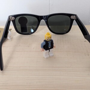 May include: Black sunglasses with dark lenses and a classic frame design. The sunglasses are open, with the arms extended. A small Lego figure stands between the lenses. A tag with the Ray-Ban logo is visible.