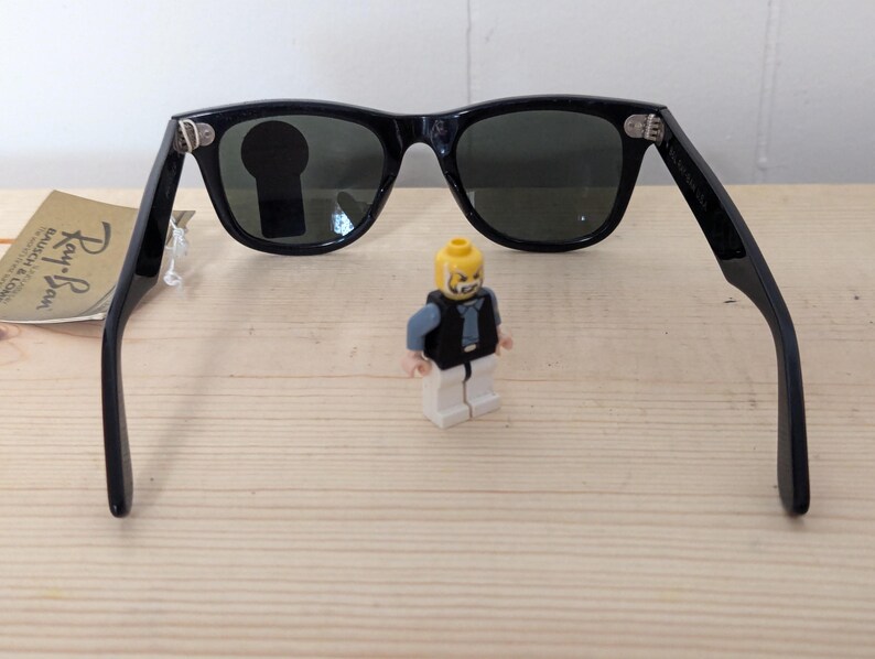 May include: Black sunglasses with dark lenses and a classic frame design. The sunglasses are open, with the arms extended. A small Lego figure stands between the lenses. A tag with the Ray-Ban logo is visible.