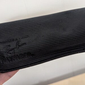 May include: Black, rectangular sunglasses case with a ribbed texture. The case has the Ray-Ban logo and the word "WAYFARER" embossed on the front. The case is held in a person's hand.