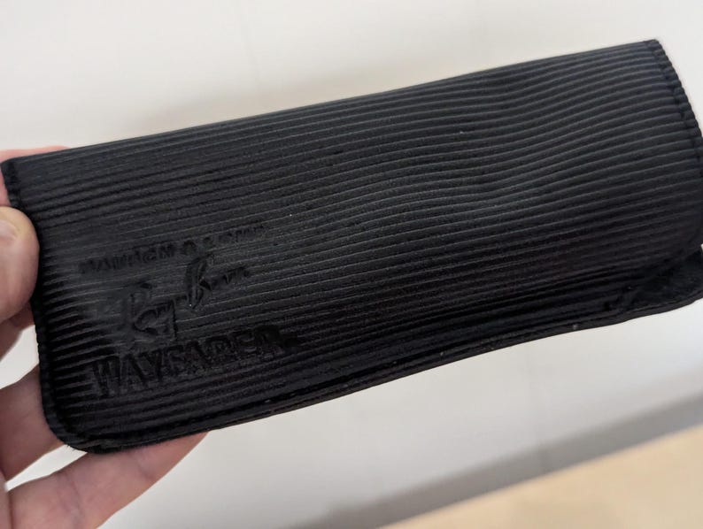 May include: Black, rectangular sunglasses case with a ribbed texture. The case has the Ray-Ban logo and the word "WAYFARER" embossed on the front. The case is held in a person's hand.