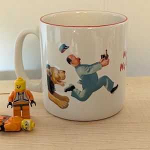 May include: White ceramic mug with a red rim featuring a cartoon illustration of a man running with a bottle, pursued by a dog. A Lego figure and pieces are in the foreground.
