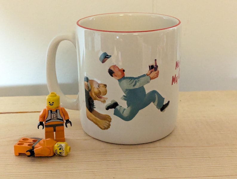 May include: White ceramic mug with a red rim featuring a cartoon illustration of a man running with a bottle, pursued by a dog. A Lego figure and pieces are in the foreground.