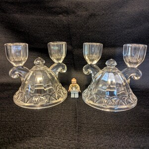May include: Two clear glass candle holders, each featuring a domed base with a textured geometric pattern and two curved arms supporting candle cups. A small plastic toy figure stands between the two glass pieces for scale comparison.