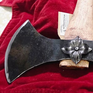Könnte beinhalten: Eine Doppelkopf-Axt mit einem dekorativen Blumenzentrum. Die Axtköpfe sind dunkel, mit einer silbernen Kante, und der Griff ist aus hellem Holz. Die Axt liegt auf einer roten Oberfläche.