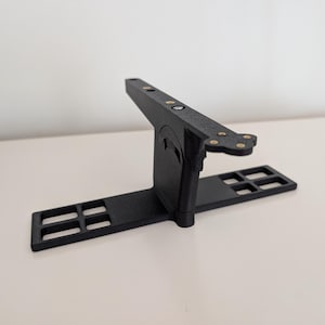 May include: A black 3D printed T-shaped stand featuring a horizontal base with four rectangular cutouts on each side. A vertical support connects to a top horizontal bar with four small circular brass-colored metal inserts embedded in the surface.