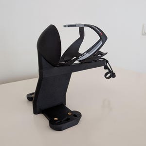 May include: A black 3D-printed bicycle water bottle holder and cage. The holder is mounted on a black base with screw holes. The cage is black with gray and white accents and holds a water bottle.