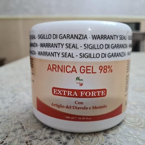 Può includere: Un barattolo bianco di Arnica Gel 98% con le parole "EXTRA FORTE" in uno striscione rosso. Il barattolo include anche il testo "Con Artiglio del Diavolo e Mentolo" e "500 ml". La parte superiore del barattolo riporta le parole "WARRANTY SEAL - SIGILLO DI GARANZIA".