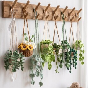 Op de afbeelding: Een verzameling handgemaakte gehaakte hangende plantenbakken, elk met een uniek ontwerp van groene bladeren en bloemen. Ze hangen aan een houten rek met pinnen, wat een charmante, handgemaakte presentatie creëert. De plantenbakken variëren in grootte en stijl.