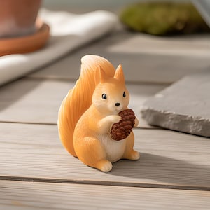 May include: A small, light orange squirrel figurine with a bushy tail, sitting upright and holding a textured brown pinecone in its front paws. The squirrel has a cream-coloured chest, pointed ears, and black eyes, presented as a decorative object.