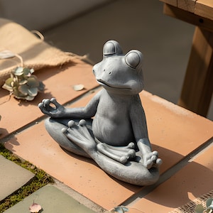 Könnte beinhalten: Eine graue Froschstatue in einer sitzenden Yoga-Pose. Der Frosch hat große Augen und sitzt im Schneidersitz mit den Händen in einer meditativen Geste. Die Statue steht auf einer Terrakotta-Fliesenoberfläche.
