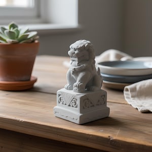 May include: A small, gray Foo Dog statue, a traditional Chinese guardian figure, sits on a decorative base. The statue is intricately detailed and placed on a wooden table. A potted succulent and tableware are in the background.