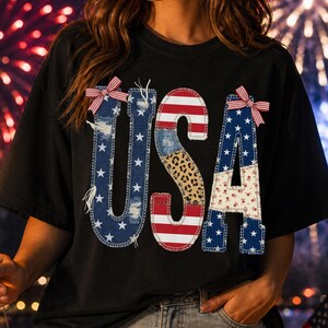 May include: Black t-shirt with the word "USA" in a patchwork design featuring stars, stripes, and leopard print. The letters are adorned with red, white, and blue bows. A person is holding a sparkler in front of a fireworks display.