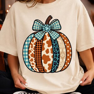 May include: A cream-colored t-shirt featuring a large graphic of a pumpkin. The pumpkin is divided into vertical segments with various patterns including blue plaid, brown plaid, cow print, leopard print, and polka dots, topped with a blue bow.