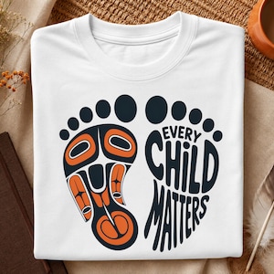 Op de afbeelding: Wit T-shirt met een grafisch ontwerp met een gestileerde voet met oranje en zwarte details, en de woorden "EVERY CHILD MATTERS" in een bijpassend lettertype. Het shirt is netjes op een getextureerd oppervlak gevouwen.