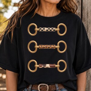 May include: A black short-sleeve t-shirt featuring a graphic print of three horizontal horse bits. The top bit has a black and white checkered pattern, the middle bit has a brown leopard print, and the bottom bit has a brown and white cow print.