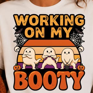 May include: A white t-shirt featuring a graphic with the text "WORKING ON MY BOOTY" in orange letters. The design includes three cartoon ghosts exercising, two jack-o'-lanterns, spider webs, bats, and skulls against a sunset background.