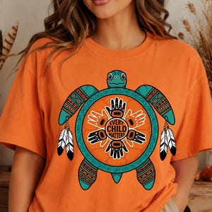May include: Orange t-shirt featuring a teal turtle design with intricate patterns and the text "EVERY CHILD MATTERS" in a circular emblem. The turtle has feathers hanging from its flippers. The shirt is a comfortable fit.