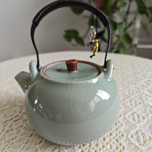 Può includere: Una teiera in ceramica verde chiaro con finitura smaltata a crepa, corpo arrotondato e beccuccio corto. Presenta un manico oscillante scuro e avvolto, decorato con due piccoli ciondoli metallici e un coperchio abbinato con pomello marrone.