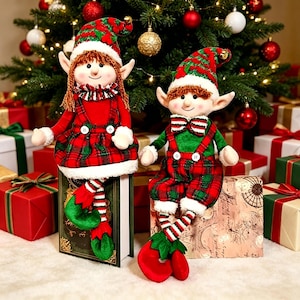 Puede incluir: Dos muñecos de duendes navideños con trajes, sombreros y zapatos a cuadros rojos y verdes. Uno lleva un vestido rojo, el otro tirantes verdes. Están sentados frente a un árbol de Navidad decorado y regalos envueltos.