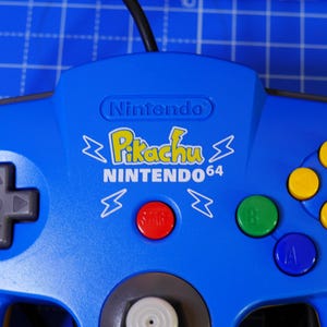 Puede incluir: Un mando de videojuegos Nintendo 64 azul con un logotipo amarillo de Pikachu, el texto Nintendo 64 y gráficos de rayos. El mando incluye una cruceta gris, un botón Start rojo, un botón B verde, un botón A azul y cuatro botones C amarillos.